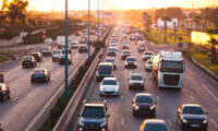 A wide view of multiple lanes filled with cars and trucks during rush hour, with a warm golden glow from the setting sun in the background.