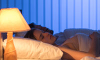 A man and woman sleep in bed with a glowing bedside lamp turned on, casting a warm light in a dark blue-lit room.