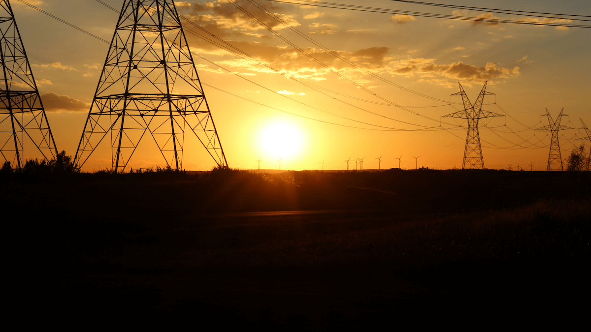 Photo of high-voltage power lines and wind turbines silhouetted against an orange sunset sky, emphasizing energy infrastructure across a flat rural landscape.
