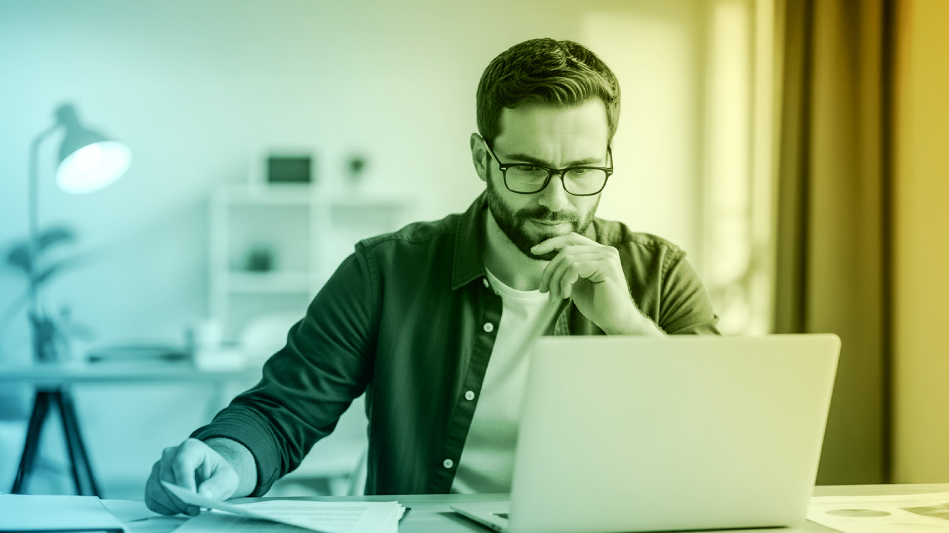A man wearing glasses and a dark button-up shirt sits at a desk, looking intently at a laptop while holding a sheet of paper. The background shows a home office with shelves and a lamp, and the image has a blue-to-yellow gradient overlay.