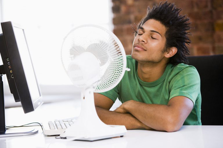 A person sitting at a desk with eyes closed, enjoying the cool air from a fan beside a computer.