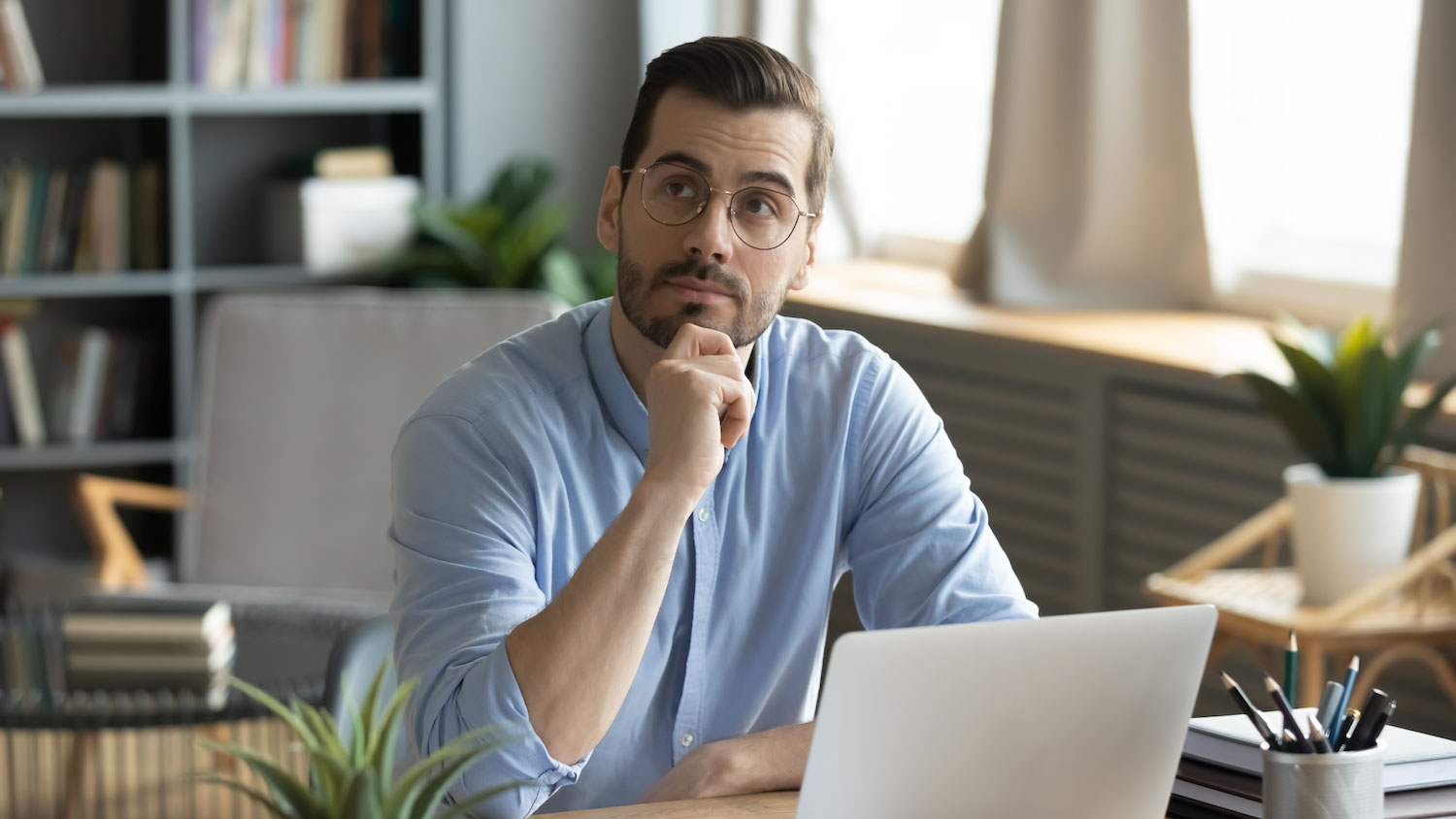 A man sitting at a desk with his hand resting on his chin, appearing thoughtful while looking upward. He sits in front of an open laptop in a bright home office with bookshelves and plants in the background. 