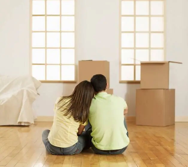 A couple sits on the floor of an empty room with their backs to the camera, leaning against each other while looking at stacked moving boxes in their new home.