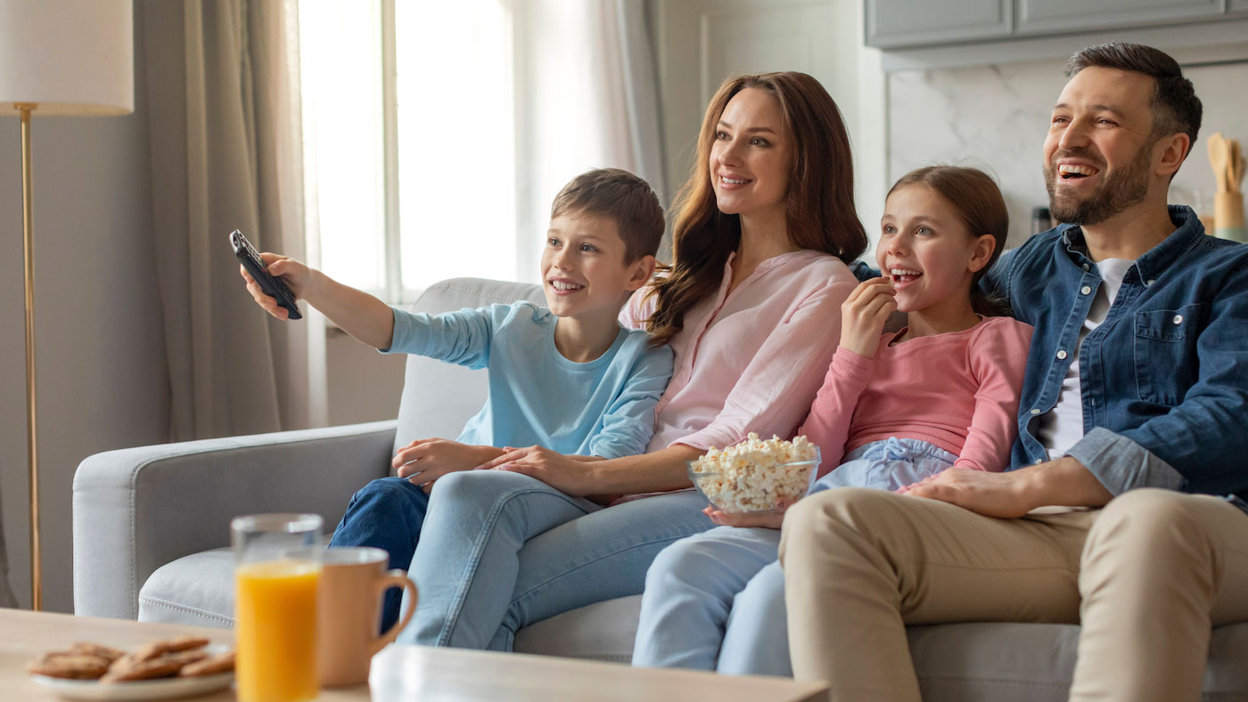 Smiling family of four sitting together on a couch, watching TV and eating popcorn in a bright living room.