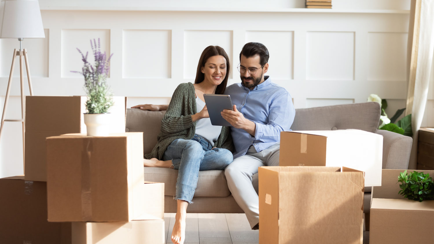 Smiling couple sitting on a couch surrounded by moving boxes, looking at a tablet together in their new home.