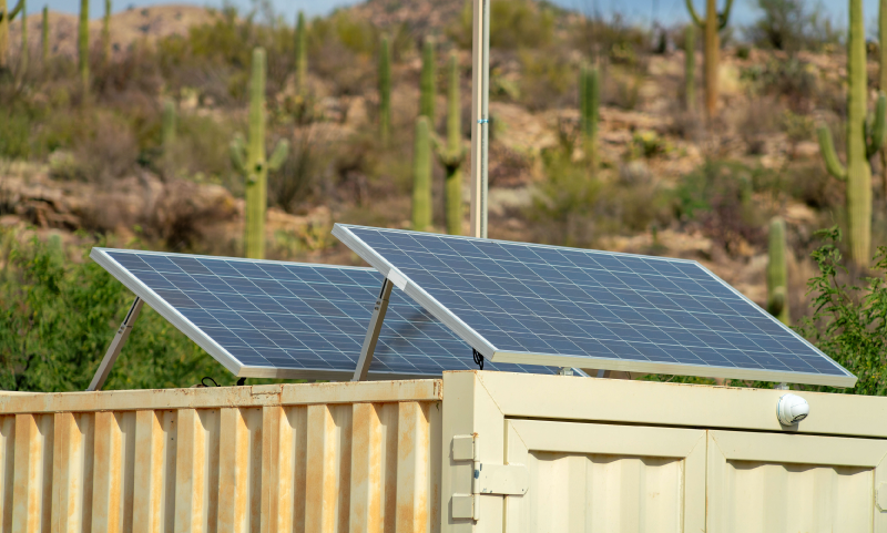 Two solar panels are mounted on a beige structure in a desert landscape with cactus plants and dry terrain in the background.