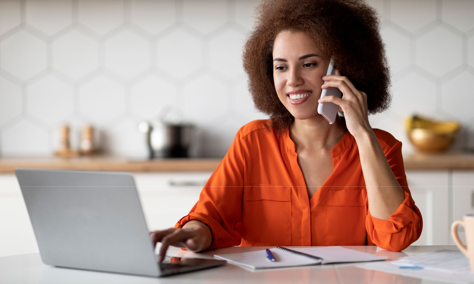Woman wearing an orange blouse sitting in a modern kitchen, smiling while talking on the phone and working on a laptop with notes on the table.