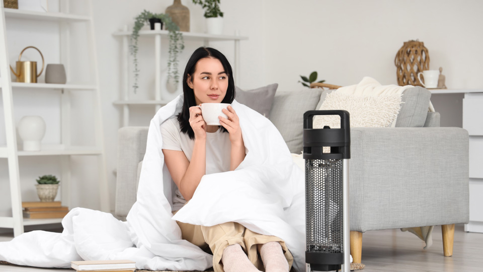 Woman wrapped in a white blanket sitting on the floor beside a portable space heater, holding a mug in a bright living room with a gray sofa.